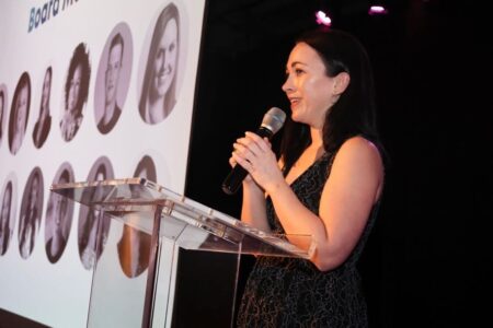 A woman in a black lace dress speaks into a microphone while standing behind a clear podium, with a screen displaying various headshot images of board members in the background, lit by purple stage lights.