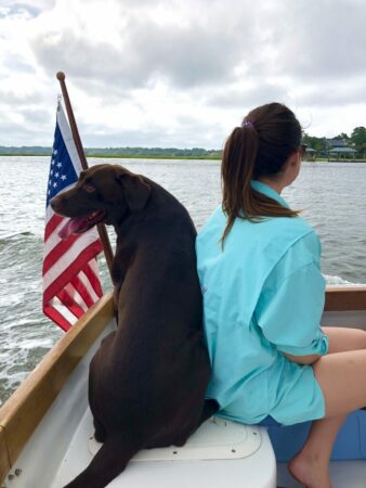 Women sitting on a boat with brown lab with American flag beside them.