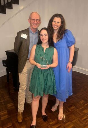 Three people standing together, with a man in a blazer and glasses on the left, a woman in a green dress holding an award in the center, and a woman in a blue dress on the right, in a well-lit room with wooden flooring and a piano in the background.