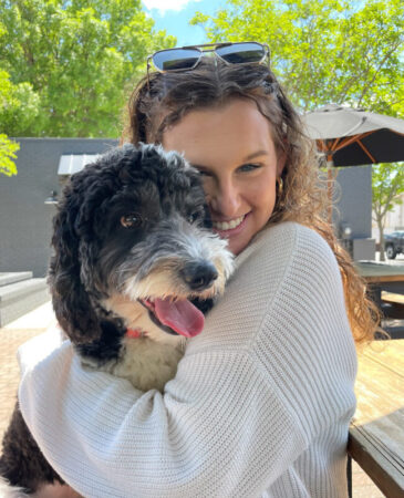 A smiling young woman with curly hair is hugging a black and white dog, enjoying a sunny outdoor setting with trees in the background and outdoor furniture visible.