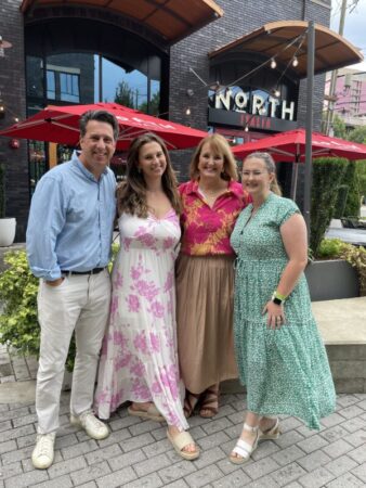 A group of four individuals poses outside a restaurant named 'North Italia.' They are standing on a brick pavement with outdoor seating in the background, featuring red umbrellas. The man on the left wears a light blue button-up shirt and white pants. The woman beside him wears a white dress with pink floral patterns. The third woman, in a pink and red floral top, stands next to the fourth woman dressed in a green patterned dress with short sleeves. All are smiling.