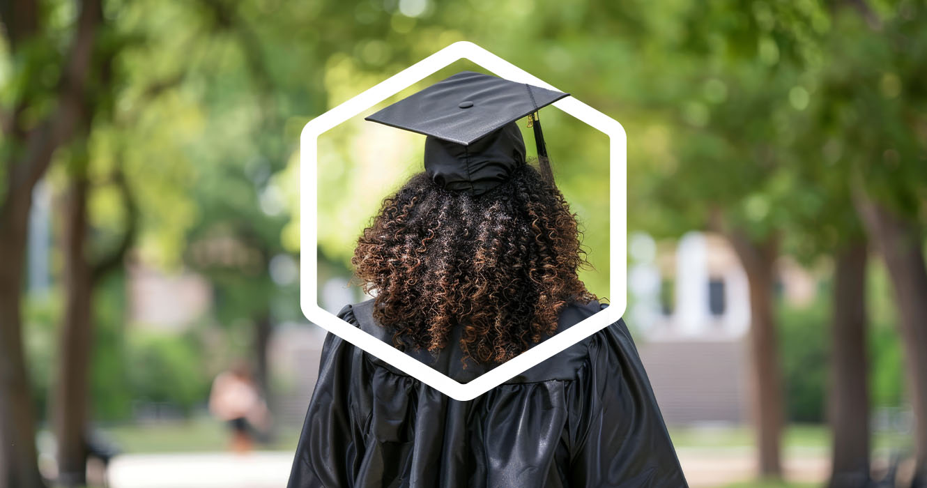 A person wearing a graduation cap and gown stands outdoors, viewed from behind, surrounded by trees.