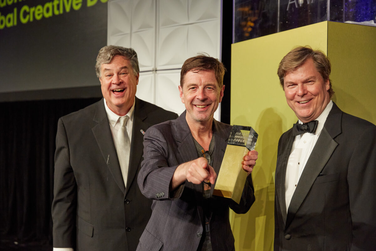 Three men in formal attire stand together on stage, with one man in the center holding an award and pointing at the camera, while the others smile. The background features a large screen displaying text and a decorated display.