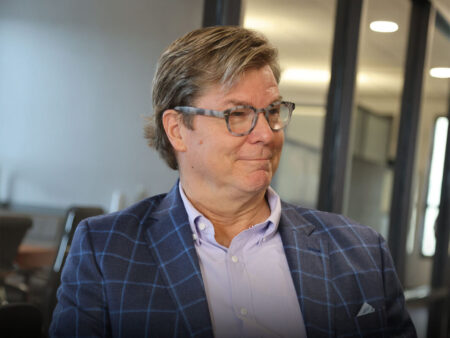 Photo of David Campbell. He is a man with light brown hair and glasses is wearing a blue plaid blazer over a light purple shirt. He is seated and appears to be smiling slightly, with a blurred office background.