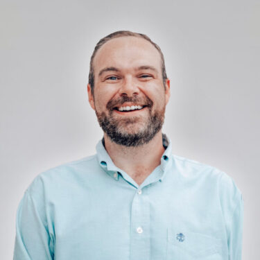 A smiling man with a beard wearing a light blue button-up shirt, set against a neutral gray background.