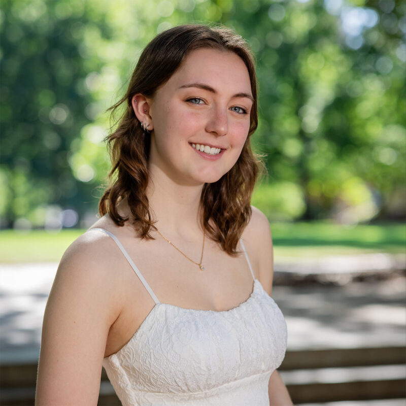 A young woman with shoulder-length wavy brown hair smiles at the camera, wearing a white dress with thin straps. She is standing outdoors against a backdrop of blurred green trees and sunlight.