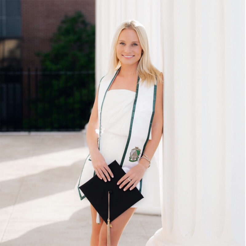 A smiling young woman in a white dress holding a black graduation cap and standing beside a large white column. She is wearing a graduation sash with green accents.