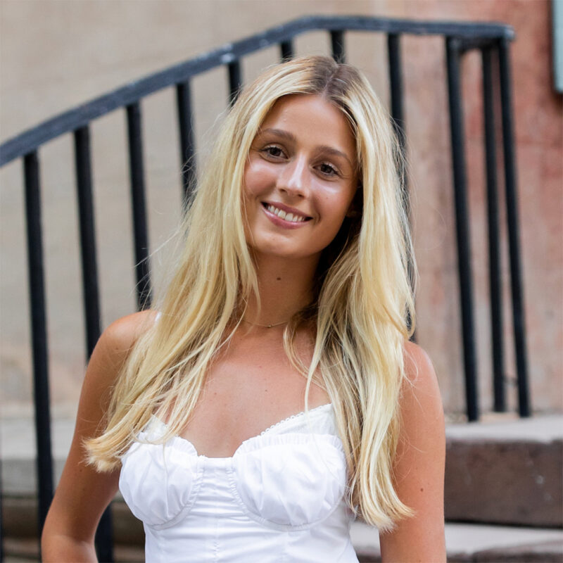 A young woman with long blonde hair smiles while standing on outdoor steps with a black railing in the background, wearing a white dress with thin straps and a sweetheart neckline.