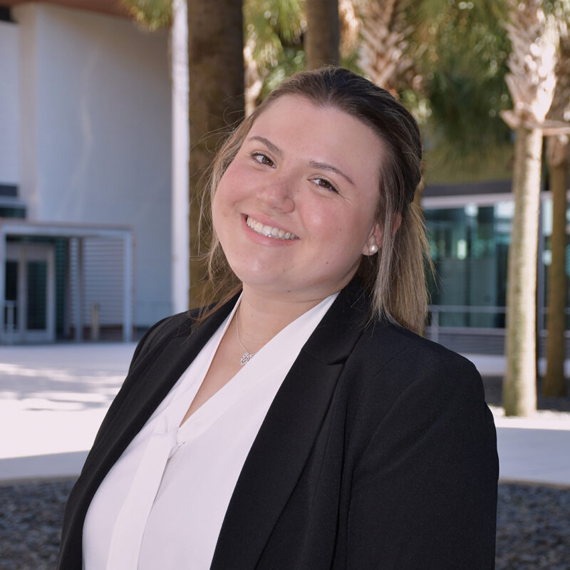A young woman with shoulder-length light brown hair smiles warmly while wearing a black blazer over a white blouse. She is standing outdoors with palm trees in the background and a modern building visible behind her.