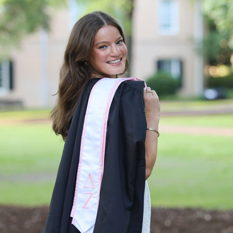 A young woman with long brown hair smiles and looks over her shoulder while wearing a black graduation gown and a white sash with pink trim, standing in a grassy area with a building in the background.