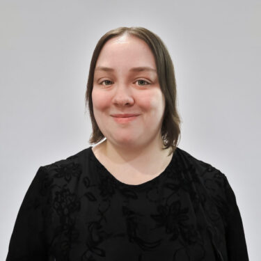 A young woman with medium-length brown hair and green eyes, wearing a black floral-patterned top, smiles at the camera against a light gray background.