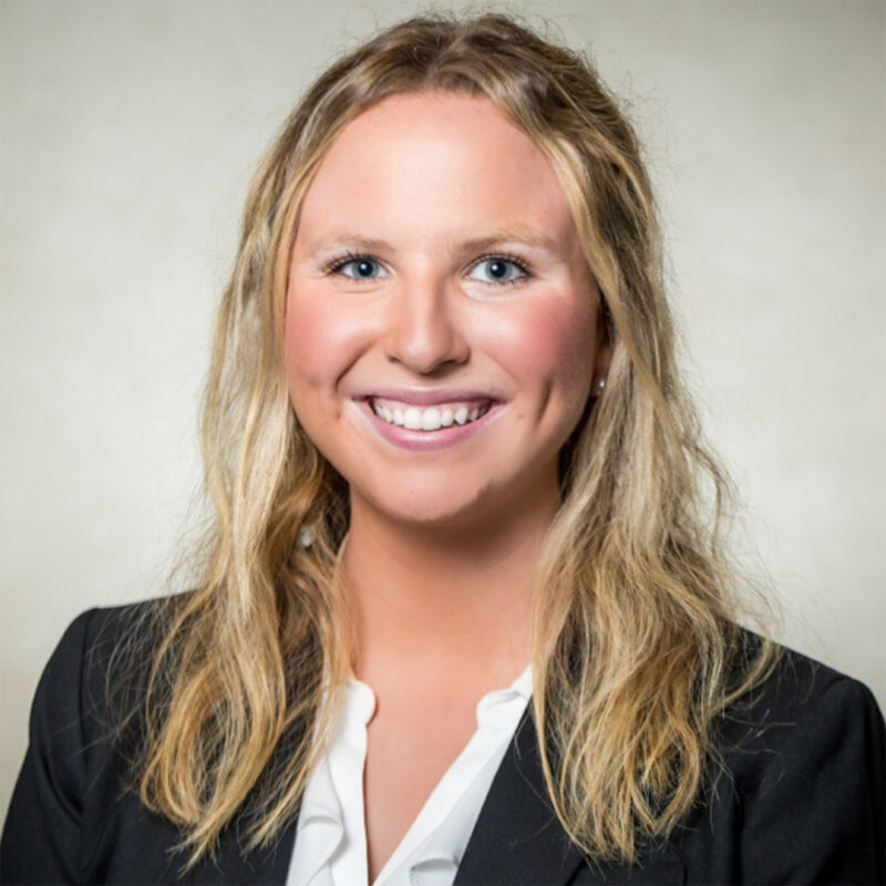 A smiling young woman with long, wavy blonde hair is wearing a black blazer over a white blouse. She is posed in front of a light, textured background.