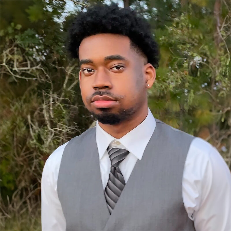 A young man wearing a gray suit vest and a white dress shirt, standing outdoors with trees in the background.