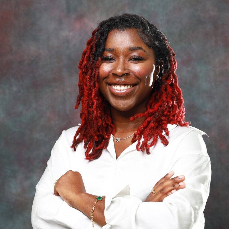 A smiling young woman with red and black dreadlocks stands confidently with her arms crossed, wearing a white blouse. The background features a textured, muted gradient.