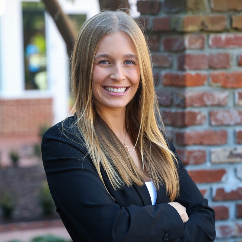 A smiling young woman with long straight blonde hair, wearing a black blazer over a white top, stands with her arms crossed in front of a brick wall and greenery in the background.