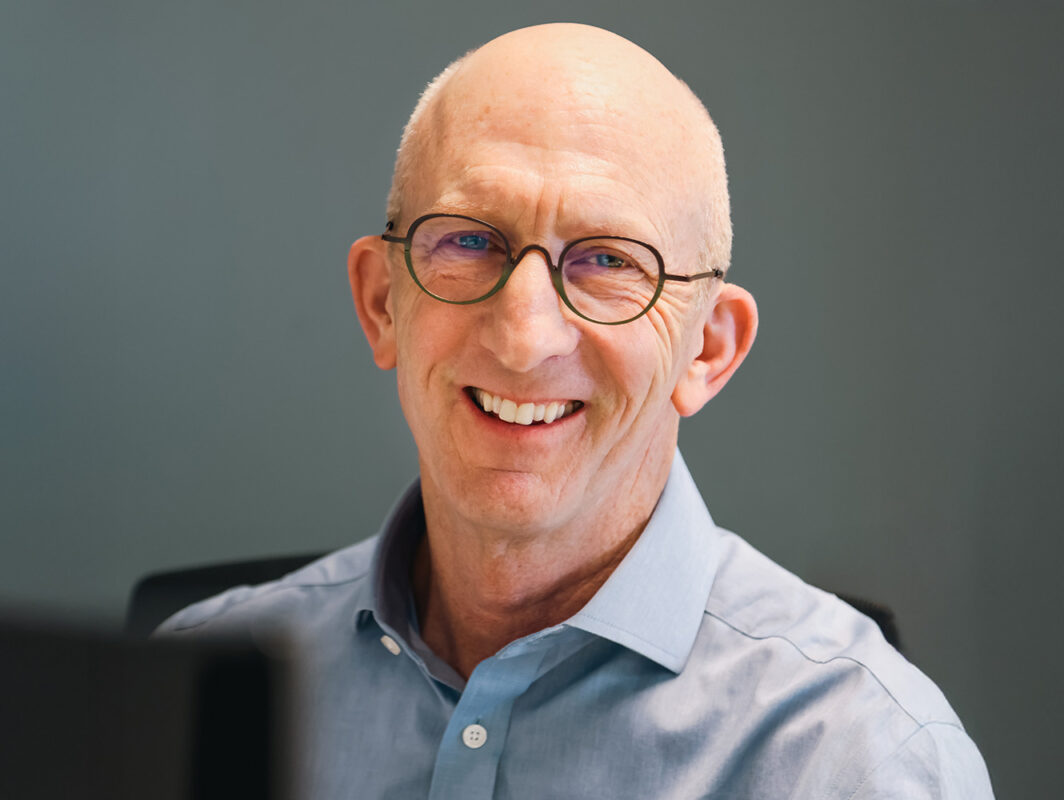 A smiling man with a bald head and glasses, wearing a light blue shirt, is sitting in an office setting with a neutral gray background.