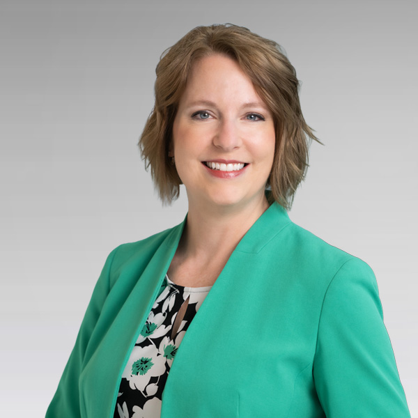 A woman with short, light brown hair smiles while wearing a green blazer over a floral-print shirt, standing against a gray background.
