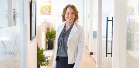 A woman in a white blazer smiles while standing in a bright, modern office corridor with plants and artwork visible.