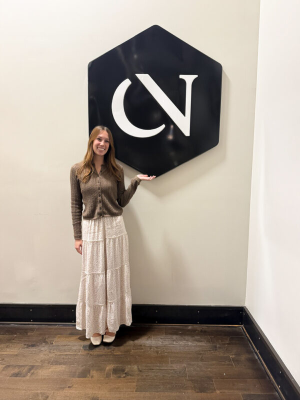 A woman stands next to a large hexagonal logo featuring the letters "CV" while wearing a brown cardigan and a long white skirt.