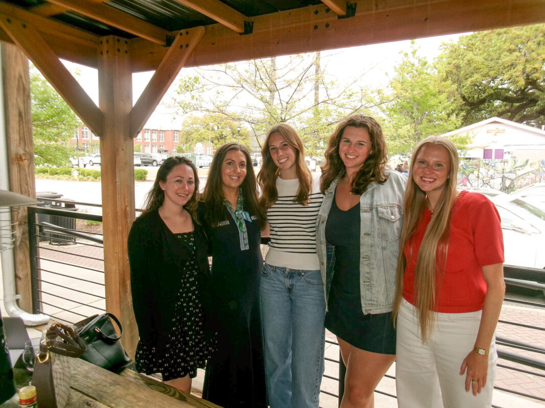 Five women stand together outdoors under a wooden structure, smiling. They are casually dressed, with greenery in the background.