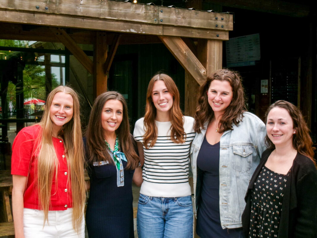 Five women stand together, smiling. They are dressed in casual attire, with different hairstyles, in an outdoor venue with wooden beams.