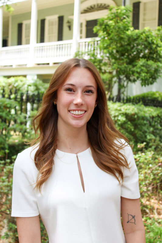 A young woman with long, wavy brown hair, wearing a white dress, smiling in front of green foliage and a historic house.