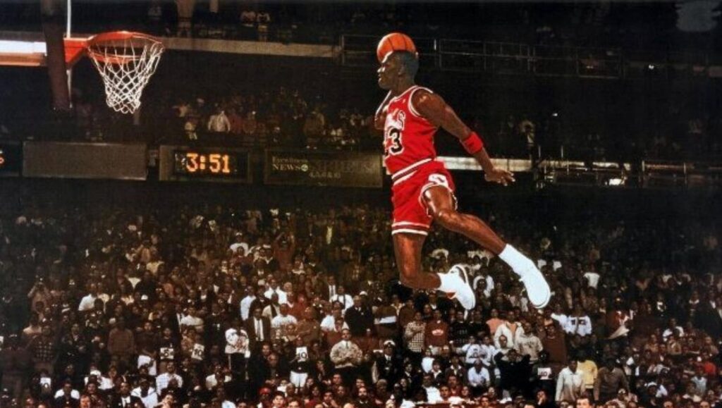 Michael Jorden with a basketball jumping toward the hoop. A large crowd in a basketball arena in the background
